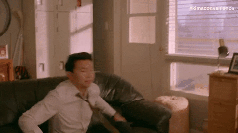 Giphy de um homem asiático sentando em um sopfá com um controle remoto na mão para assistir televisão.